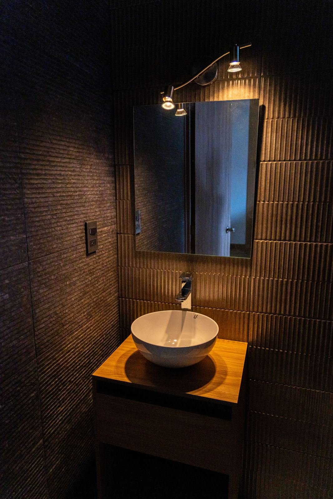 Baño con muros acanalados, ovalín sobre cubierta de madera y spots sobre el espejo