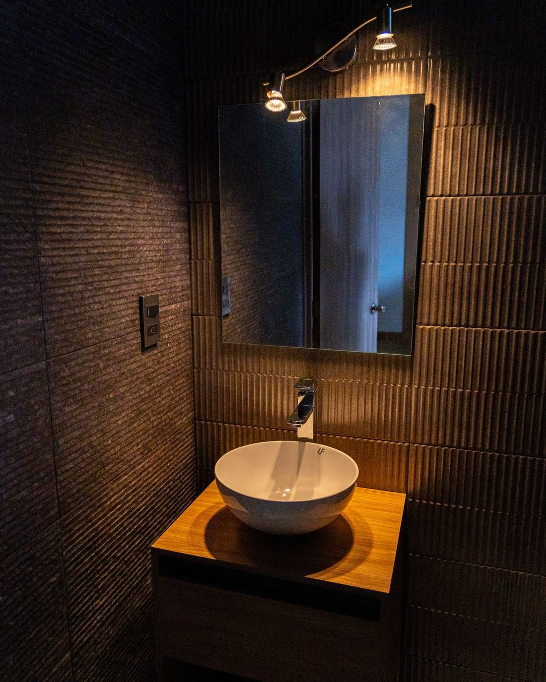 Baño de Hospital Concordia con muros acanalados, ovalín sobre mueble de madera y luz cálida
