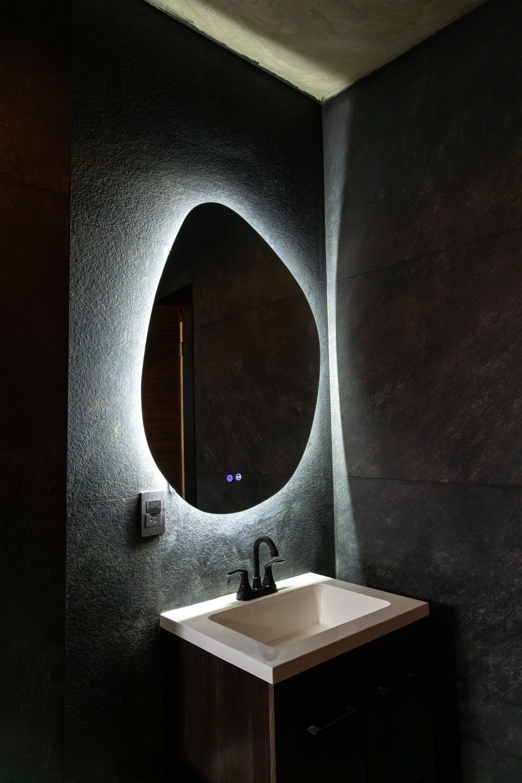 Baño con espejo orgánico retroiluminado, muro de piedra negra y lavabo con mueble oscuro
