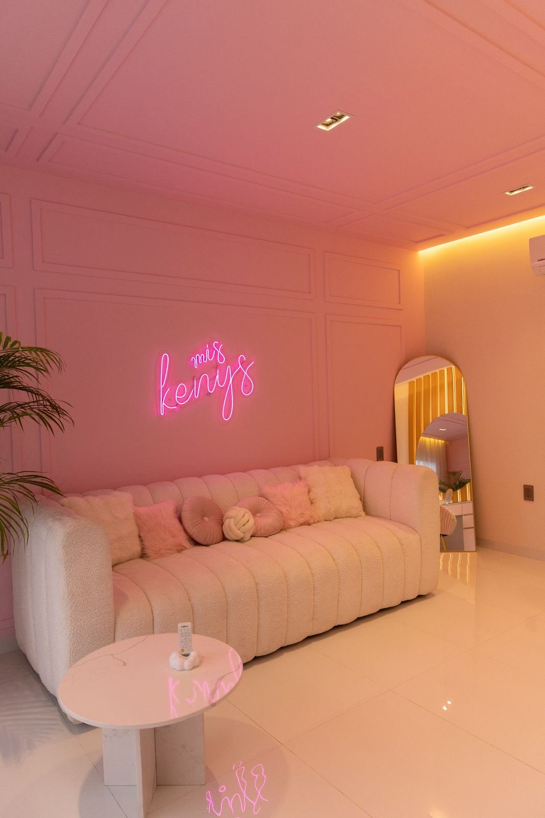 Sala con muros rosas de molduras, sofá bouclé, letrero de neón y espejo de arco